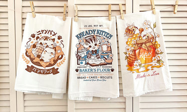 Three cotton flour sack tea towels featuring soft, vintage-style art strung up on a clothesline in front of wood slat wall door.