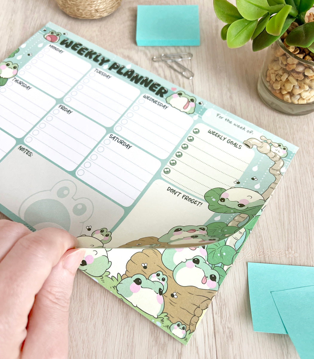A weekly planner notepad on a wood table, the print featuring a collection of fat, blob-like frogs decorating a checklist style planner page. A hand lifts the corner of the top page.