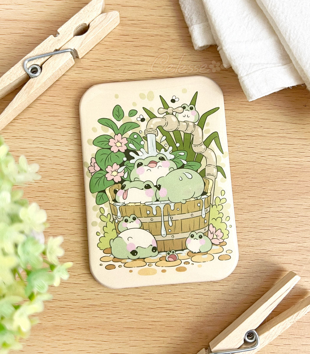 A photo of a fridge magnet laid on a table, the artwork featuring a group of blob-shaped frogs splashing around under a faucet in a wooden bucket, plants all around.