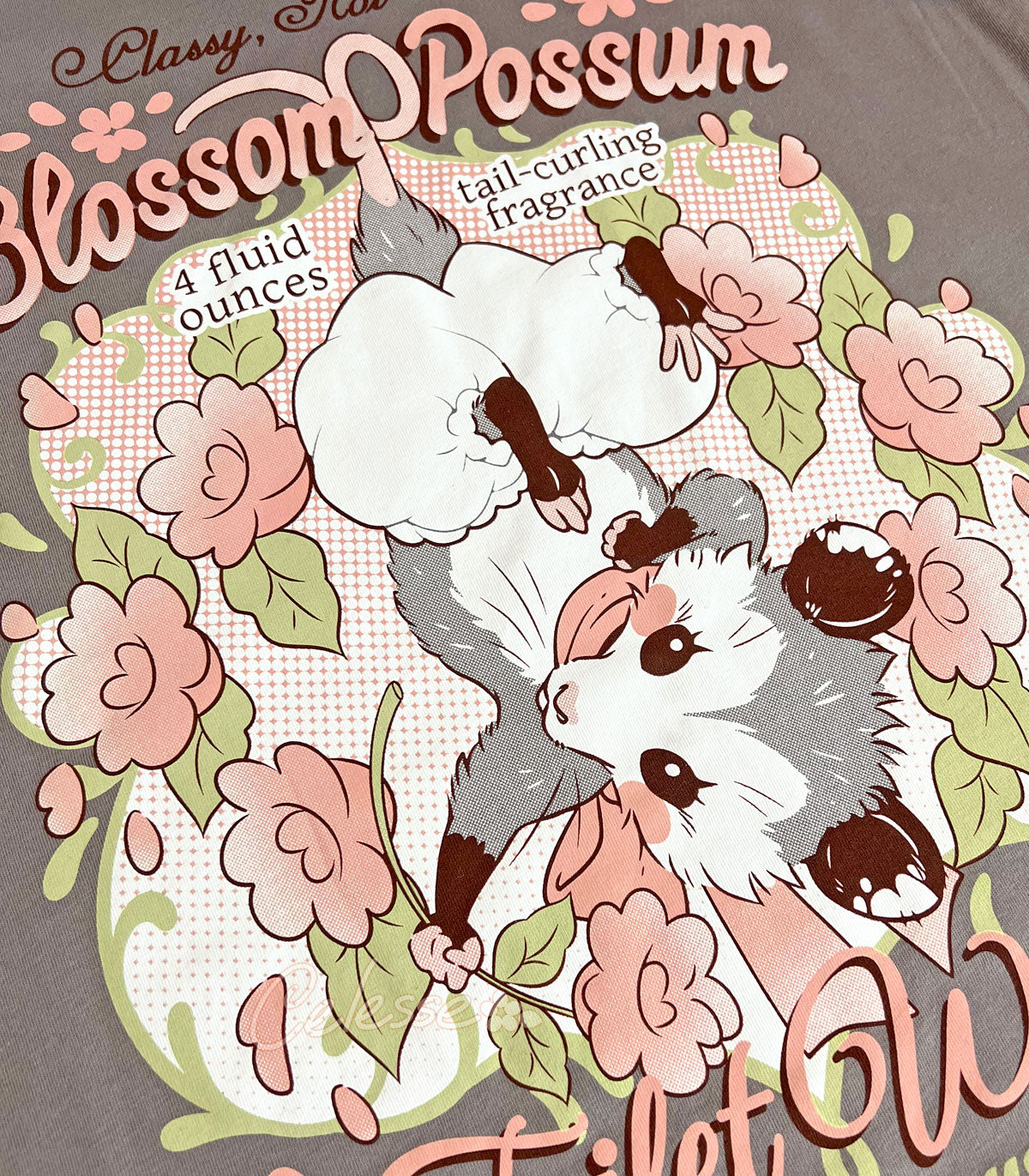 A close-up photo of the print of a warm gray shirt with a vintage-style logo featuring a hanging baby opossum surrounded by flowers.