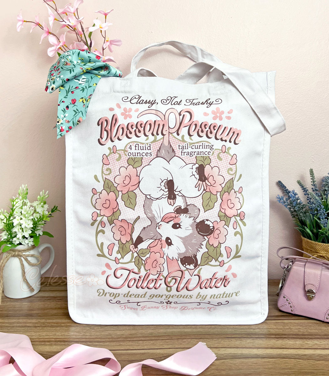 A white canvas tote bag with a whimsical vintage-style logo for Blossom Opossum Toilet Water, styled on a wooden surface with decorative flowers, a pink purse, and a floral scarf.