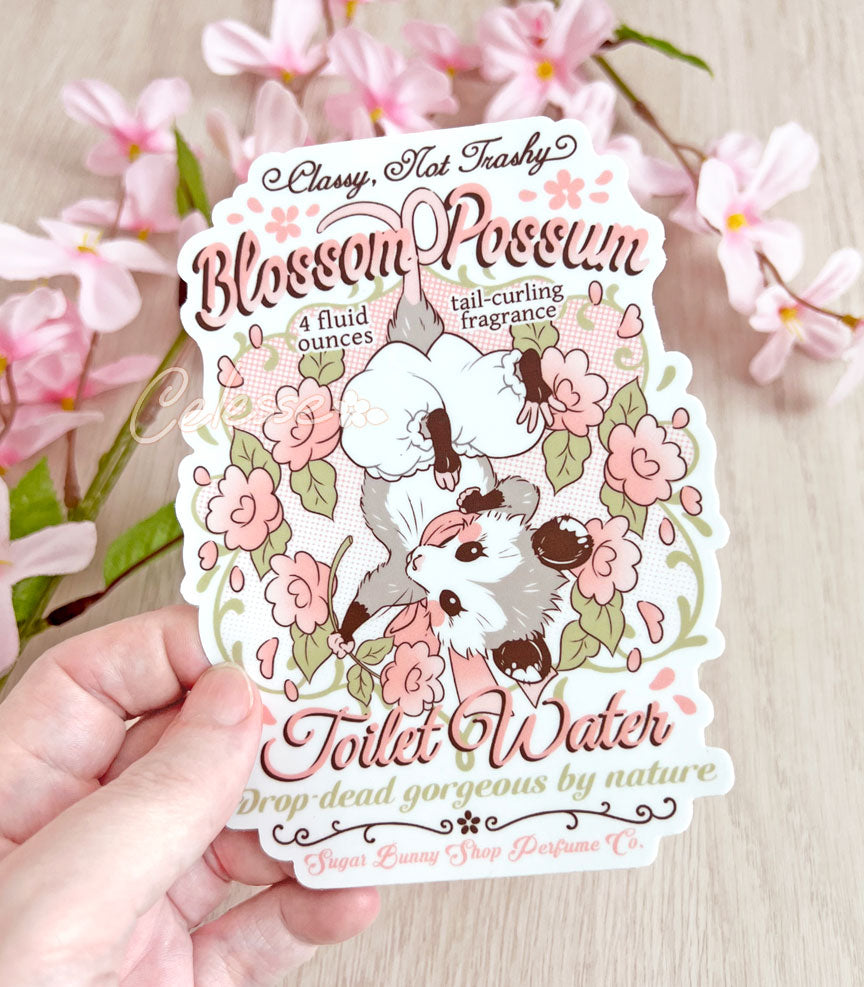 An extra large vinyl sticker of a vintage-style logo for Blossom Opossum Toilet Water, the art featuring a baby opossum in bloomers hanging upside down and surrounded by blossoms. The flavor text reads "Classy, Not Trashy" and "Drop-dead gorgeous by nature."