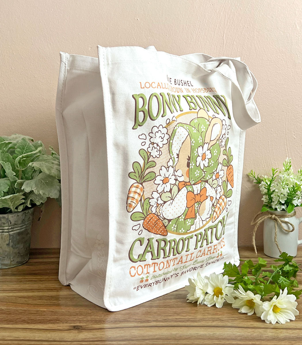 A photo of a white canvas tote on a table against a warm pink wall, plants and flowers on either side of it. The front of the bag features a print of a faux vintage-style logo for Bonny Bunny Carrot Patch, with a bunny in a bonnet holding a bundle of flowers and carrots around her.