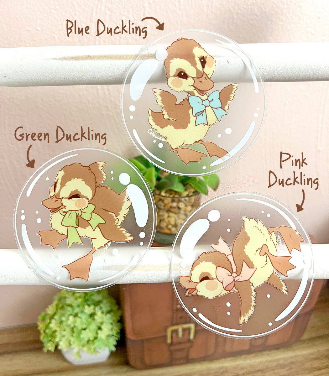 A photo of three clear vinyl stickers featuring artwork of ducklings floating inside bubbles, displayed on a white wood stand with a neutral background.