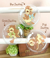 A photo of three clear vinyl stickers featuring artwork of ducklings floating inside bubbles, displayed on a white wood stand with a neutral background.