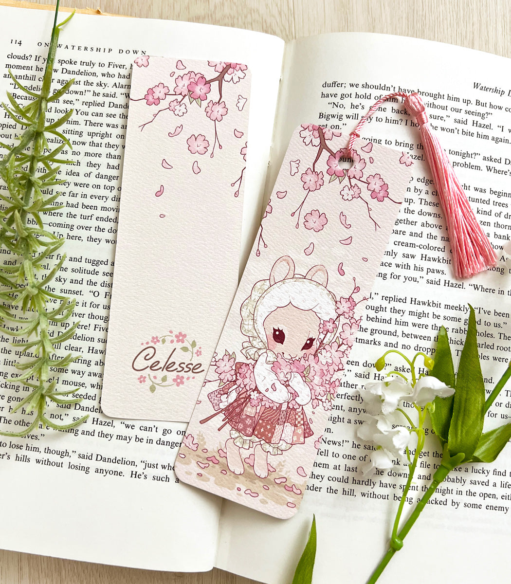 A photo of a bookmark placed atop an open book, the artwork featuring a bunny wearing an old fashioned dress and bonnet and holding a bushel of cherry blossom branches.