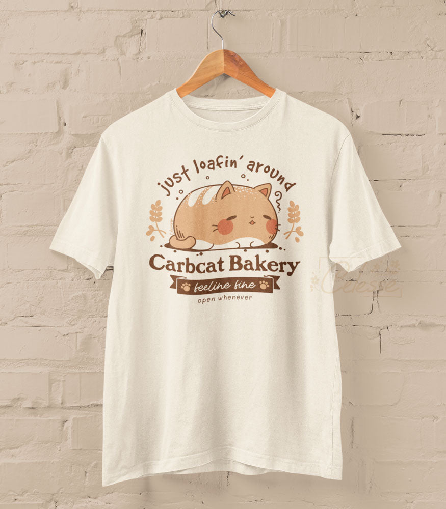 A photo of a shirt on a wood hanger set against a beige brick wall, the print featuring a fat bread-like cat and the title Carbcat Bakery. The tagline reads "just loafin&