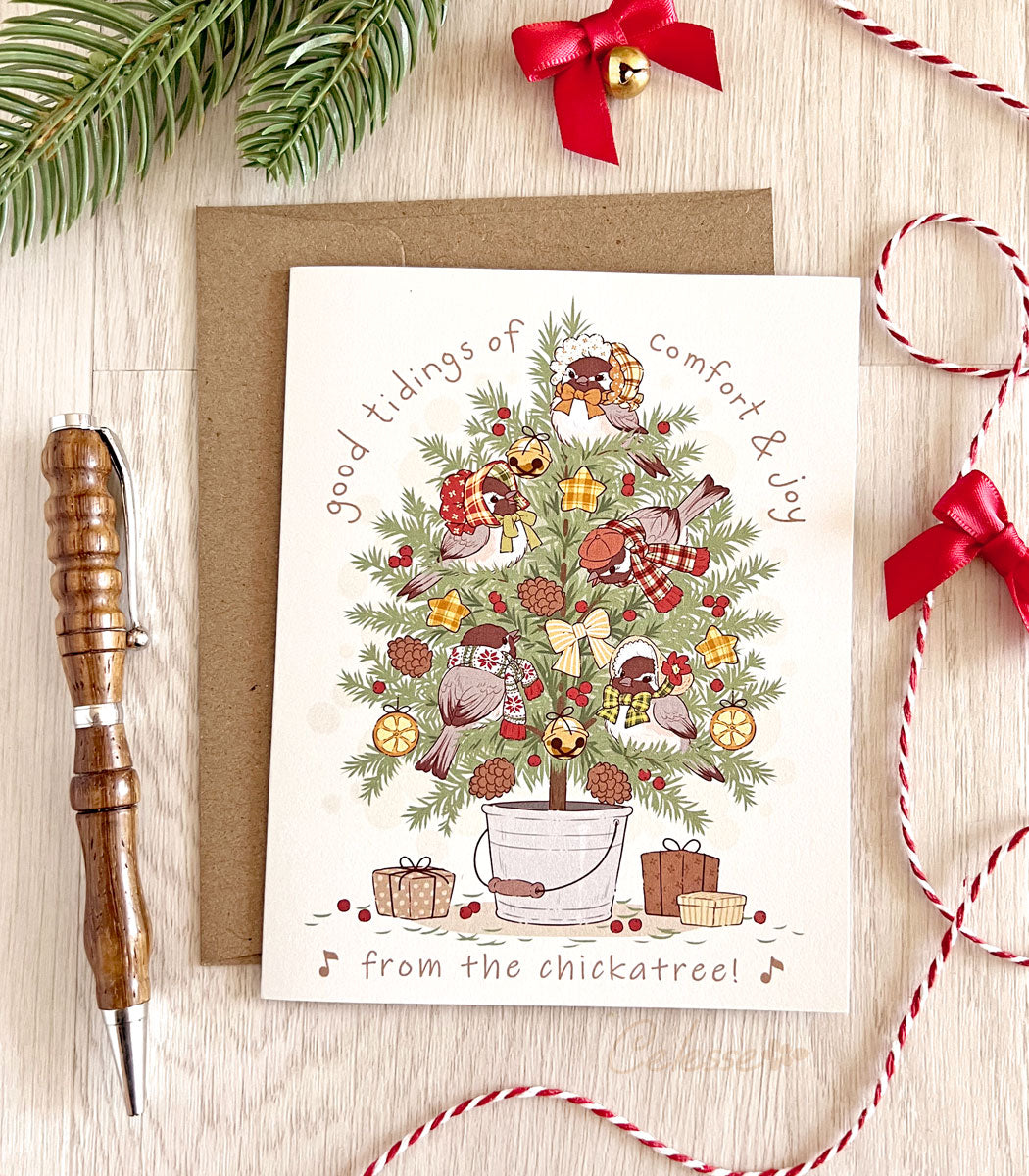 A Christmas greeting card with an illustration of chickadees in a decorated tree, accompanied by festive elements like gifts and pine branches. The card is placed next to a kraft envelope, with a red and white string and a wooden pen in the background.