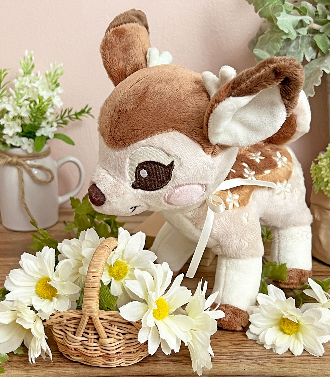 A photo of a baby deer plush on a table with a mini wicker basket of white flowers in front of them, more plants and flowers in the background. The fawn&