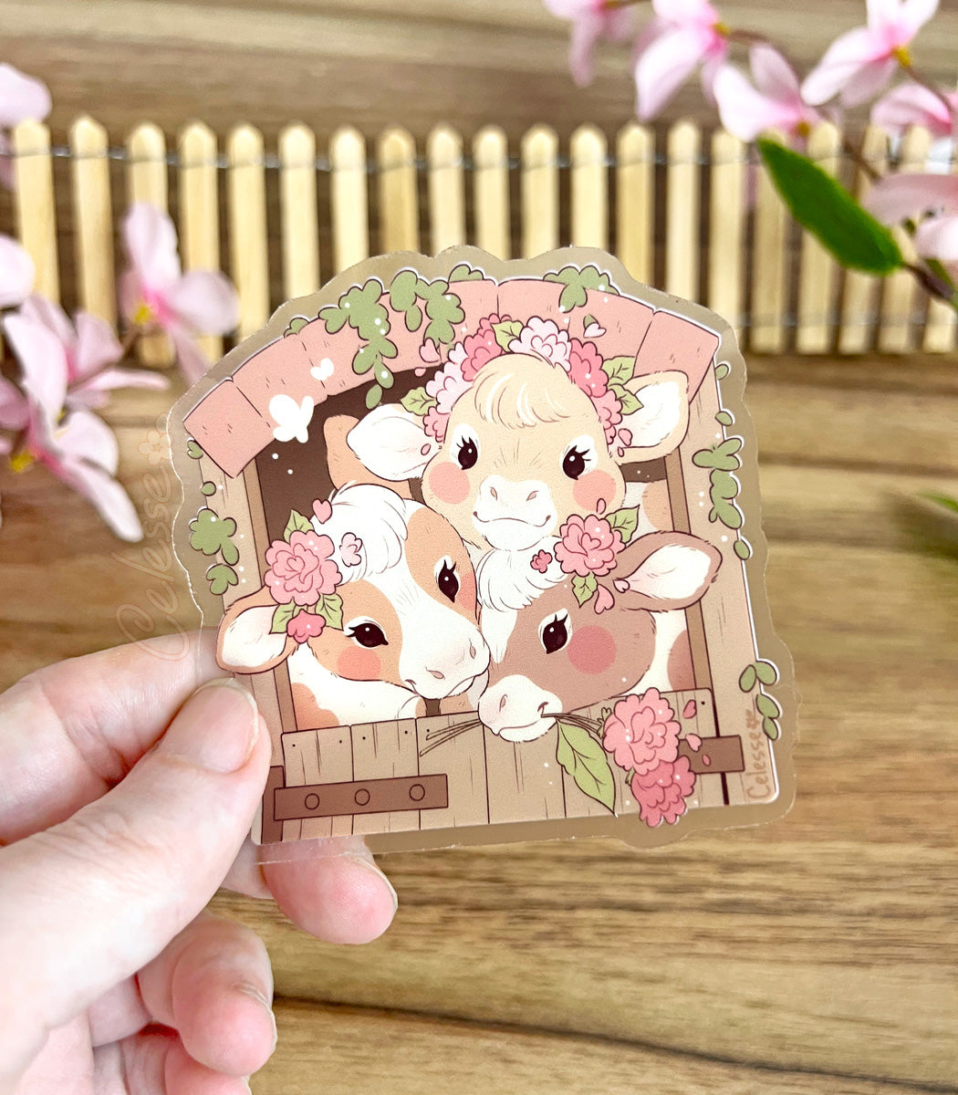 A photo of a handing holding a clear vinyl sticker with country cows in a barn above a wooden surface with flowers and a small fence in the background.