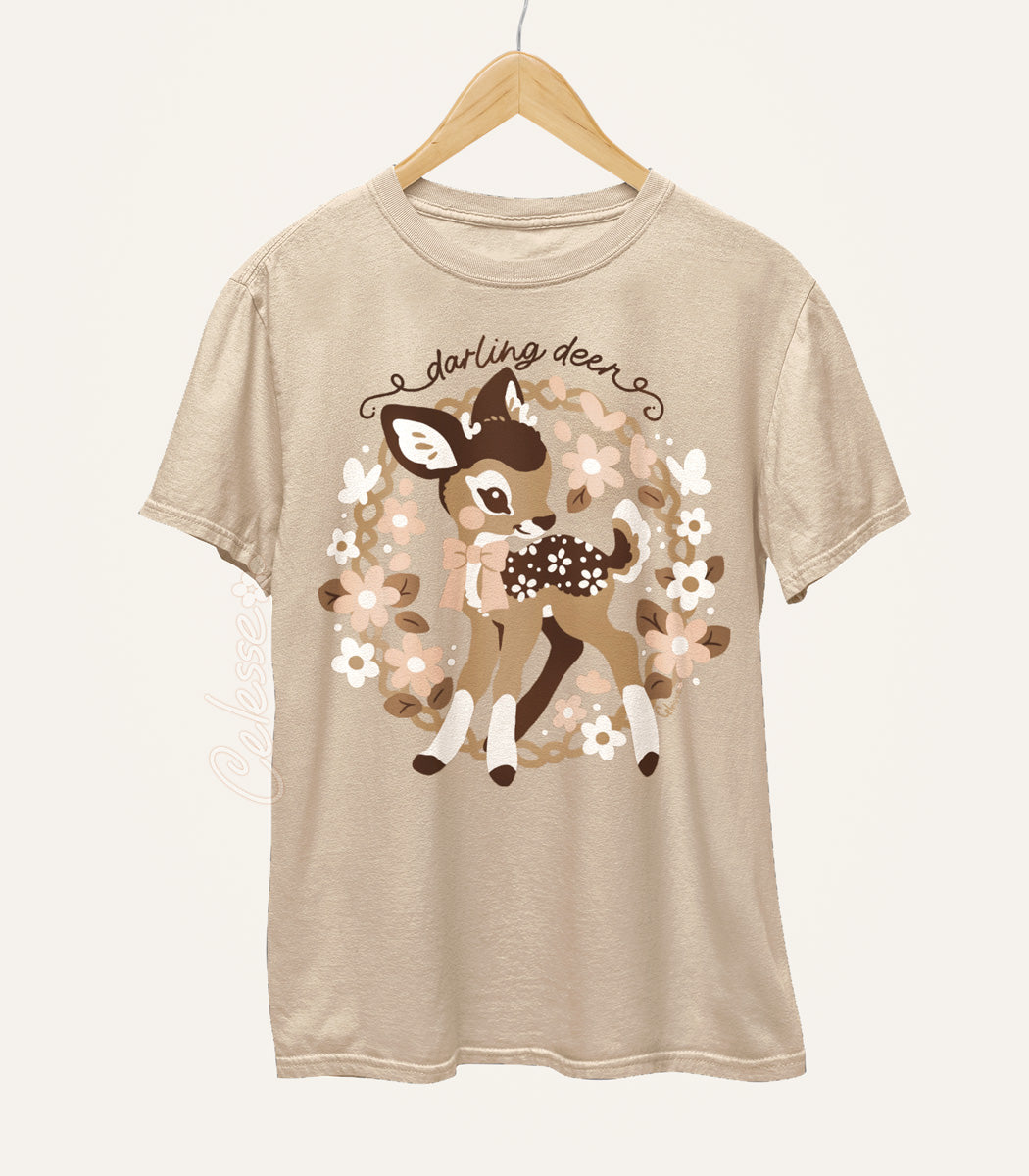 A tan-colored shirt on a wood hanger featuring a print of a baby deer with flower spots, surrounded by a wreath. The text reads "darling deer".
