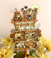 A photo of a large clear vinyl sticker perched above yellow flowers with artwork of a haunted home filled with cute little witch cats.