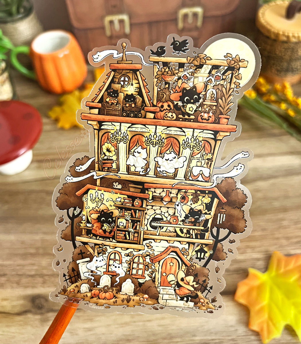 Photo of a clear vinyl sticker with artwork of a decorative Halloween-themed house full of witch cats over a wooden surface with pumpkins and leaves.