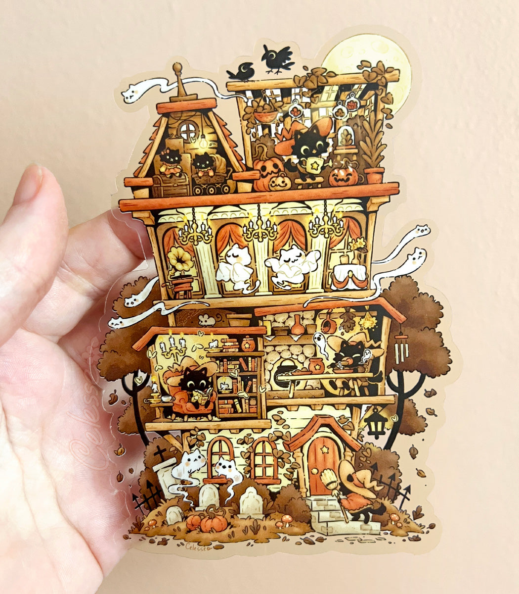 A hand holding a large clear vinyl sticker with an illustration of a haunted house with witch cats and various spooky elements on a beige background.