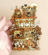 A hand holding a large clear vinyl sticker with an illustration of a haunted house with witch cats and various spooky elements on a beige background.