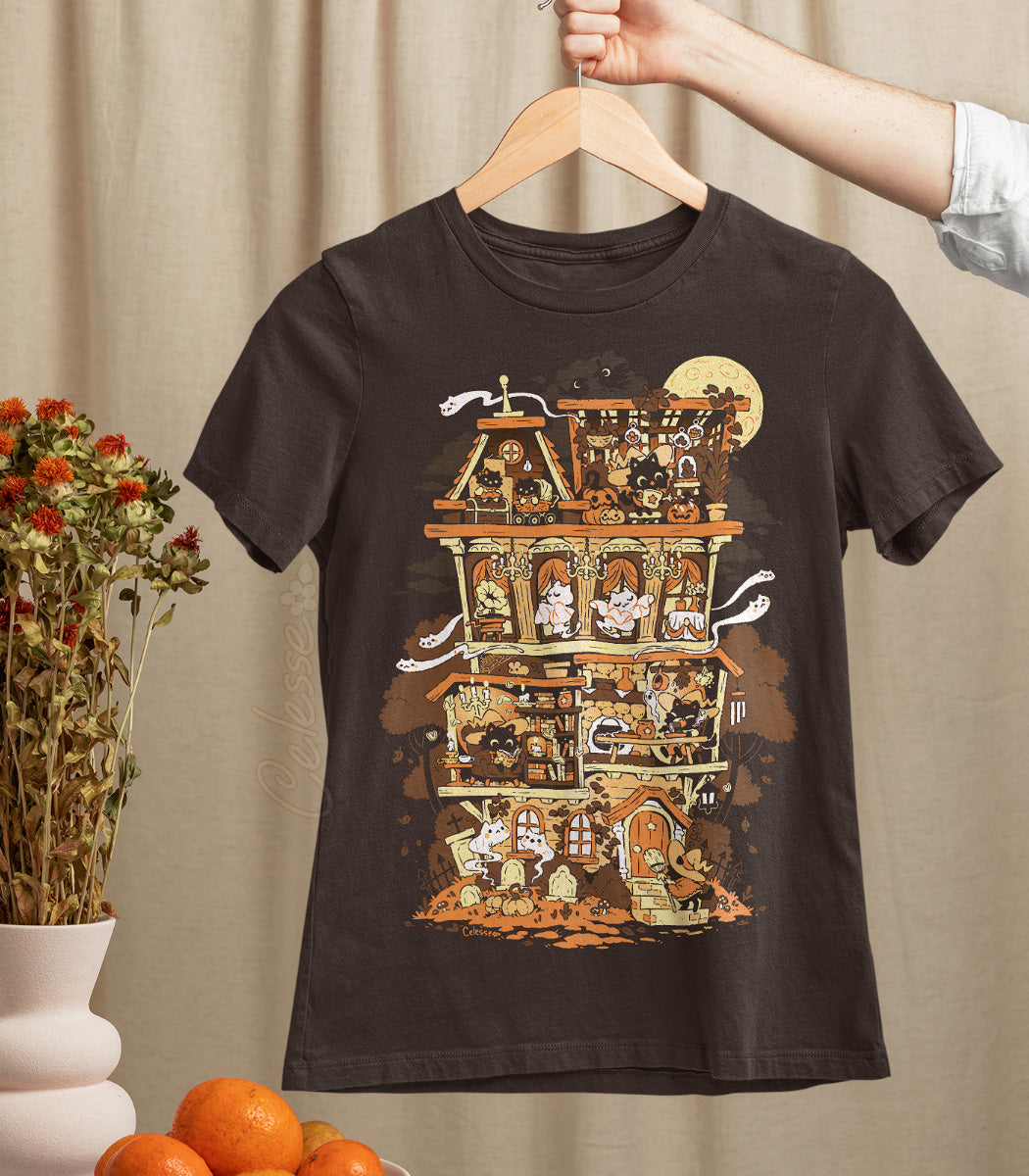 A hand holding up a dark brown shirt on a wooden hanger, the print featuring a Halloween-themed haunted home full of witch cats and ghosts.