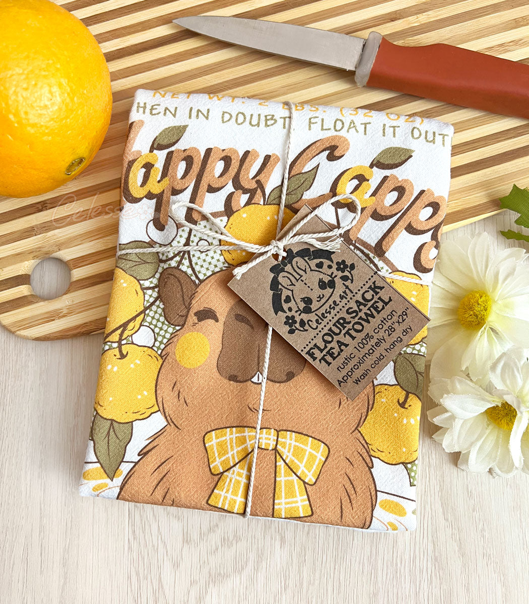 A folded kitchen tea towel tied with twine with a vintage-style logo for Happy Cappy Yuzu Fruit featuring a blissful capybara, styled on a wooden cutting board next to an orange, a small knife, and white flowers.