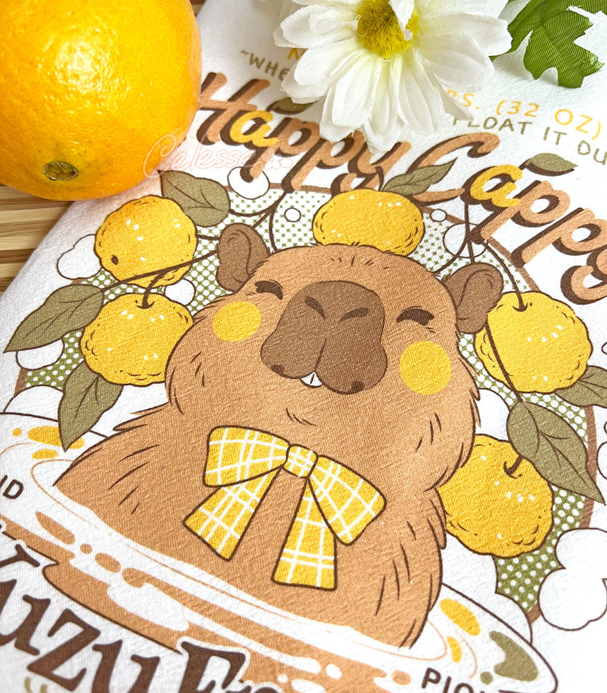 A close up of a white kitchen tea towel with a vintage-style logo for Happy Cappy Yuzu Fruit featuring a blissful capybara, styled on a wooden cutting board next to an orange and white flowers.
