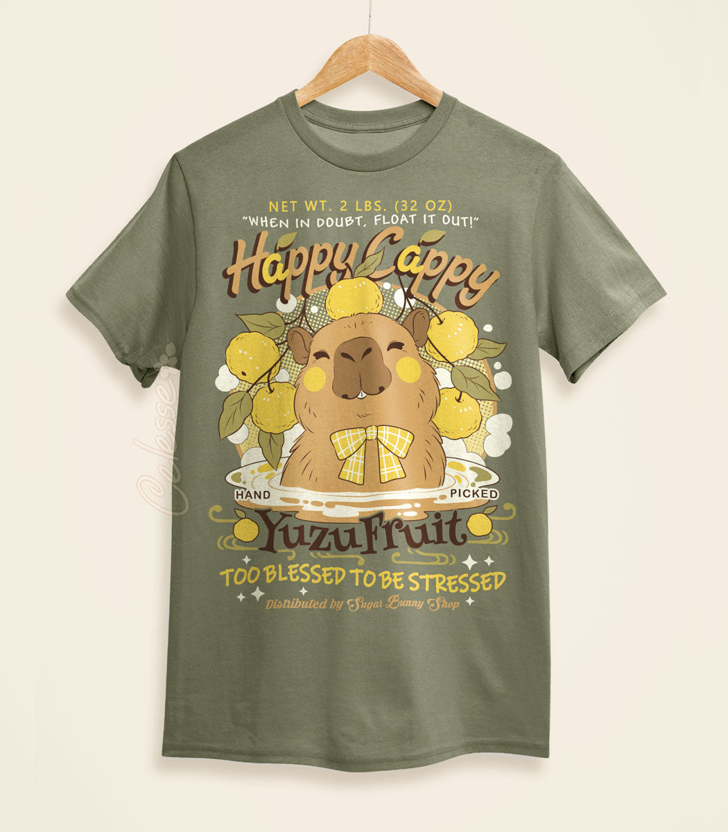 A light olive green shirt on a wood hanger with a print of a vintage-style logo for Happy Cappy Yuzu Fruit, the center art featuring a smiling capybara in a hot spring.