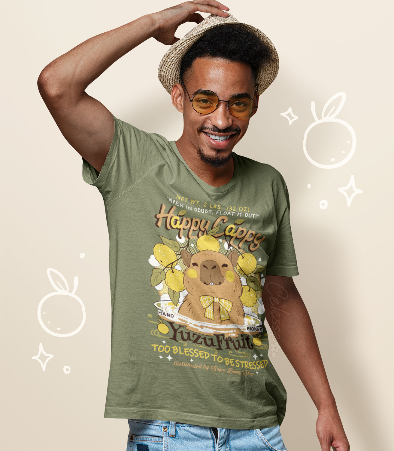 A man in a straw cap and sunglasses, wearing a light olive green shirt with a print of a vintage-style logo for Happy Cappy Yuzu Fruit, the center art featuring a smiling capybara in a hot spring.