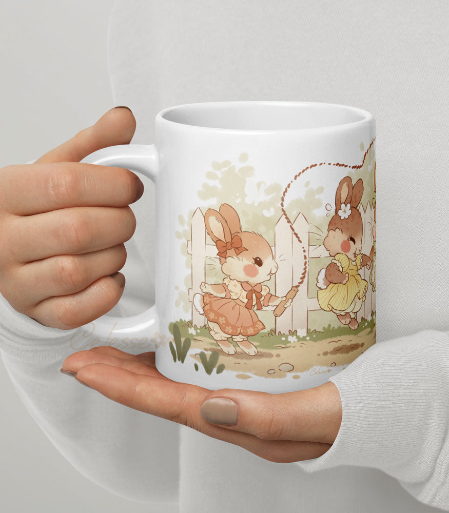 Hands holding a 20 oz. ceramic mug with a print featuring art of a bunch of bunny girls playing jump rope in vintage dresses.