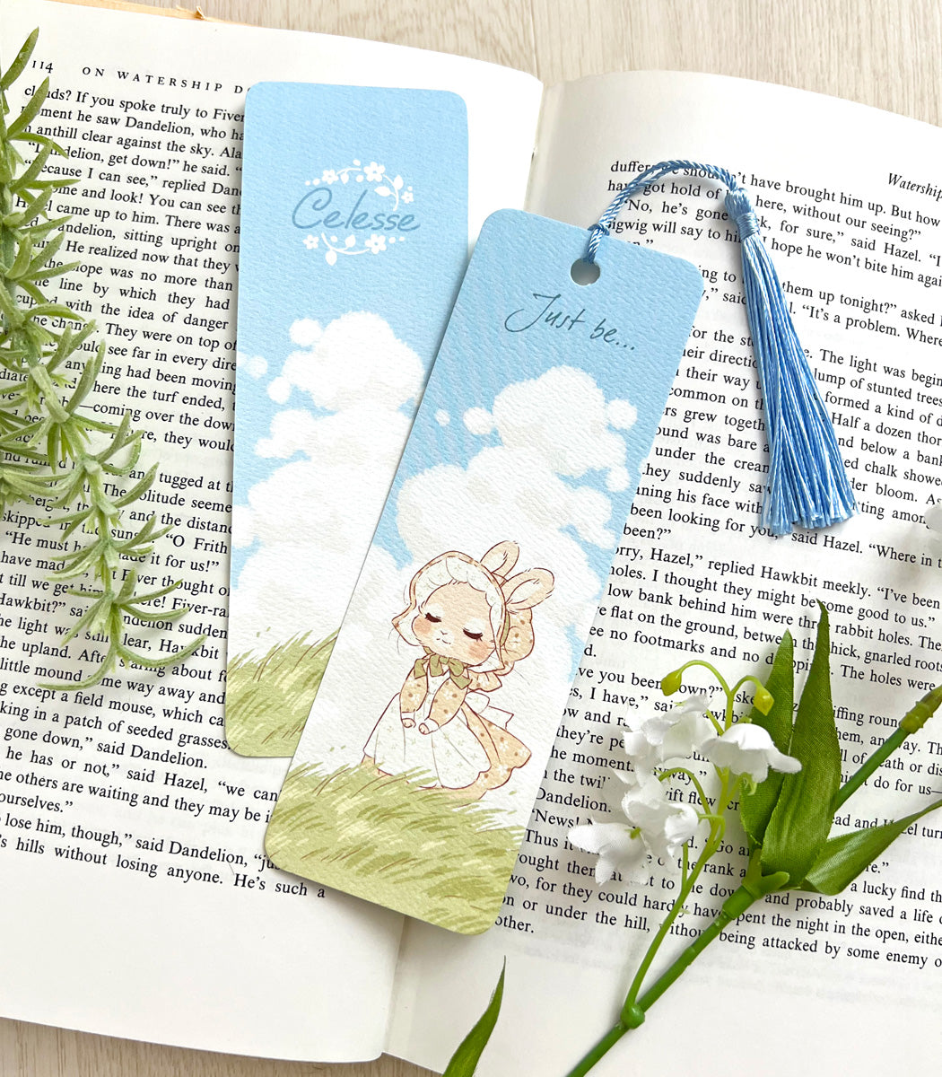 A photo of a bookmark placed atop an open book, the artwork featuring a bunny in a vintage dress and bonnet perched serenely atop a grassy hill on a sunny day while a breeze blows. The text reads "Just be..."