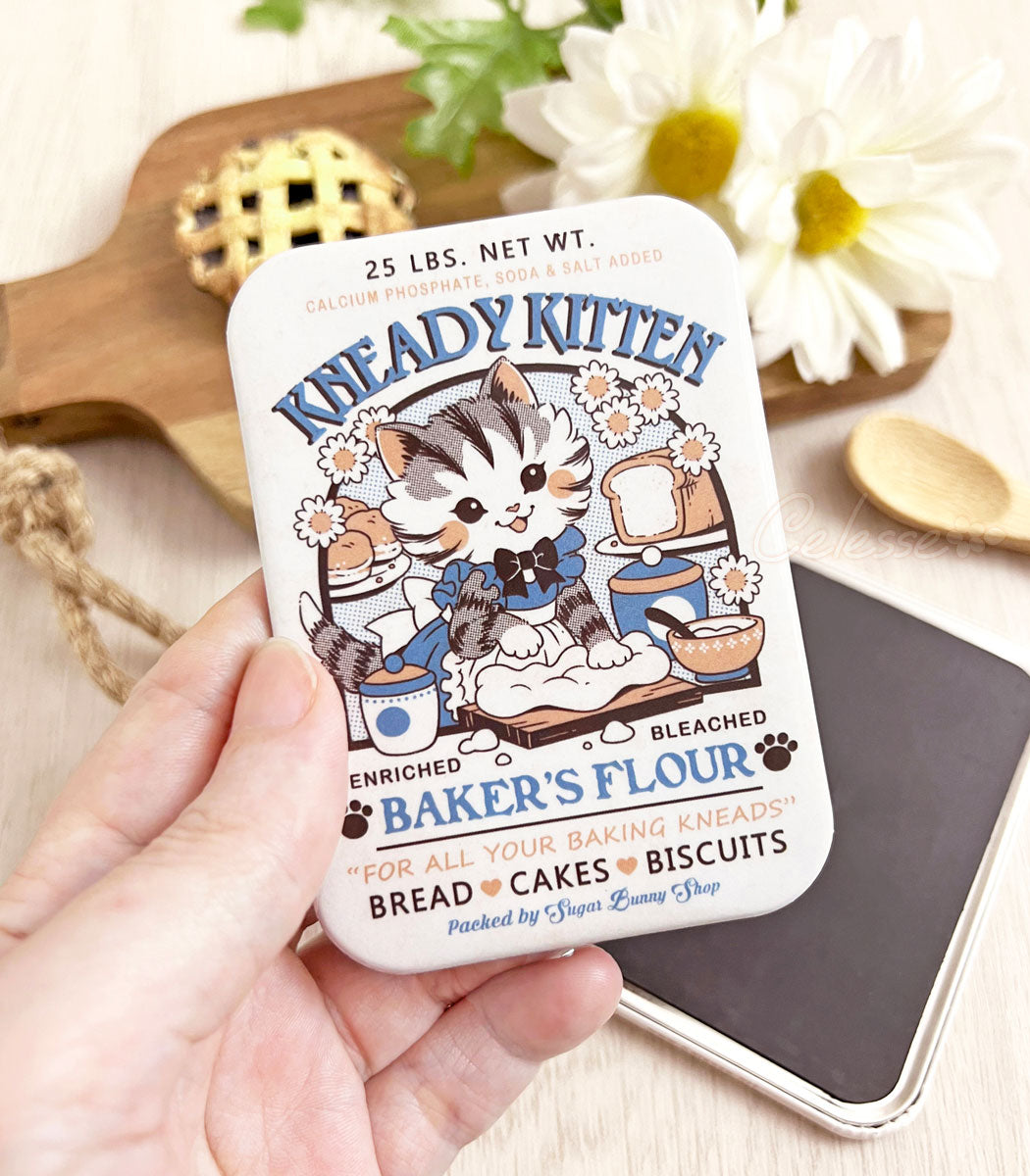 A photo of a hand holding a rectangular refrigerator magnet featuring artwork of a vintage-style logo for Kneady Kitten Baker&