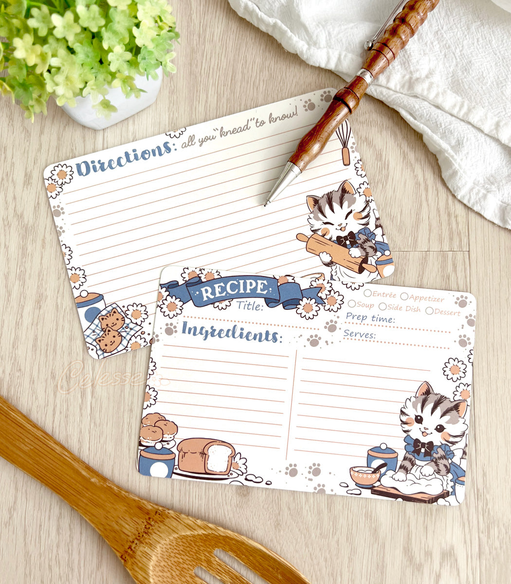 Two recipe cards with cat illustrations on a wooden surface with kitchen utensils.