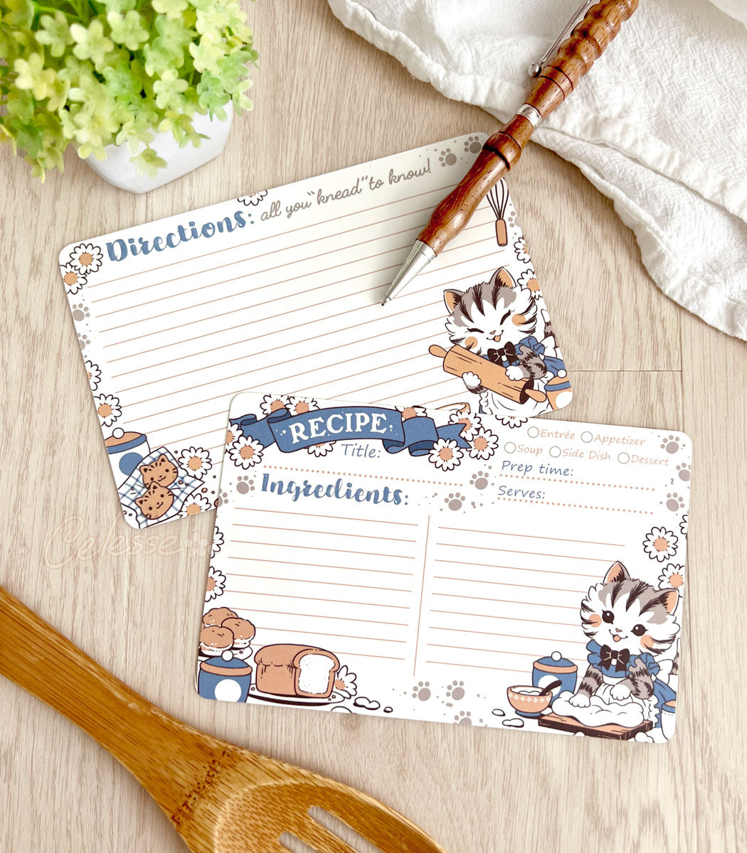 Two recipe cards showing front and back on a wooden surface with kitchen utensils, the design featuring vintage-style art of a baking kitten wearing a frilly dress and apron.
