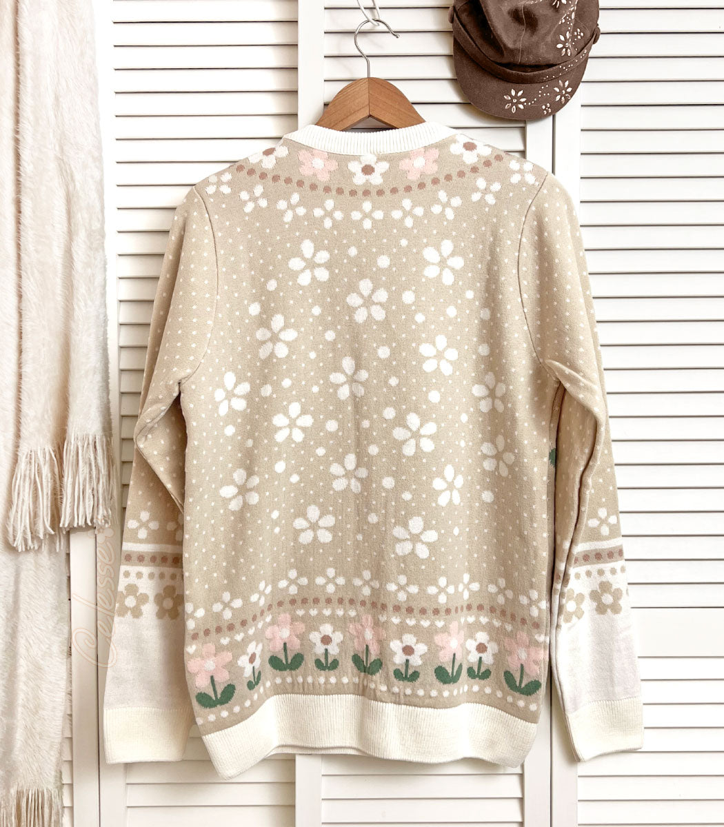 A photo of the back side of a knit sweater hung from a wood hanger on a white slat wall door. The colors are soft beige and browns and the design features a speckling of white flower shapes scattered over the back.
