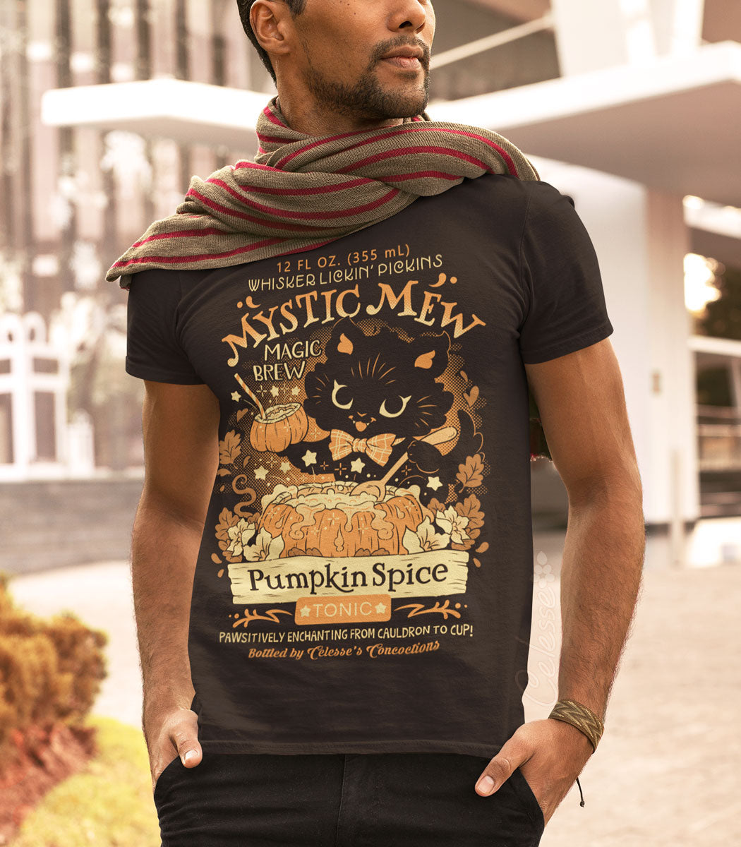 A photo of a black man standing outdoors in autumn with his hands in his pockets, wearing a striped scarf and dark brown t-shirt with a print featuring a logo for Mystic Mew Pumpkin Spice Tonic, a black kitten stirring a pumpkin cauldron full of magic brew popping with stars and sparkles and holding up a cup-sized pumpkin with a straw in it.
