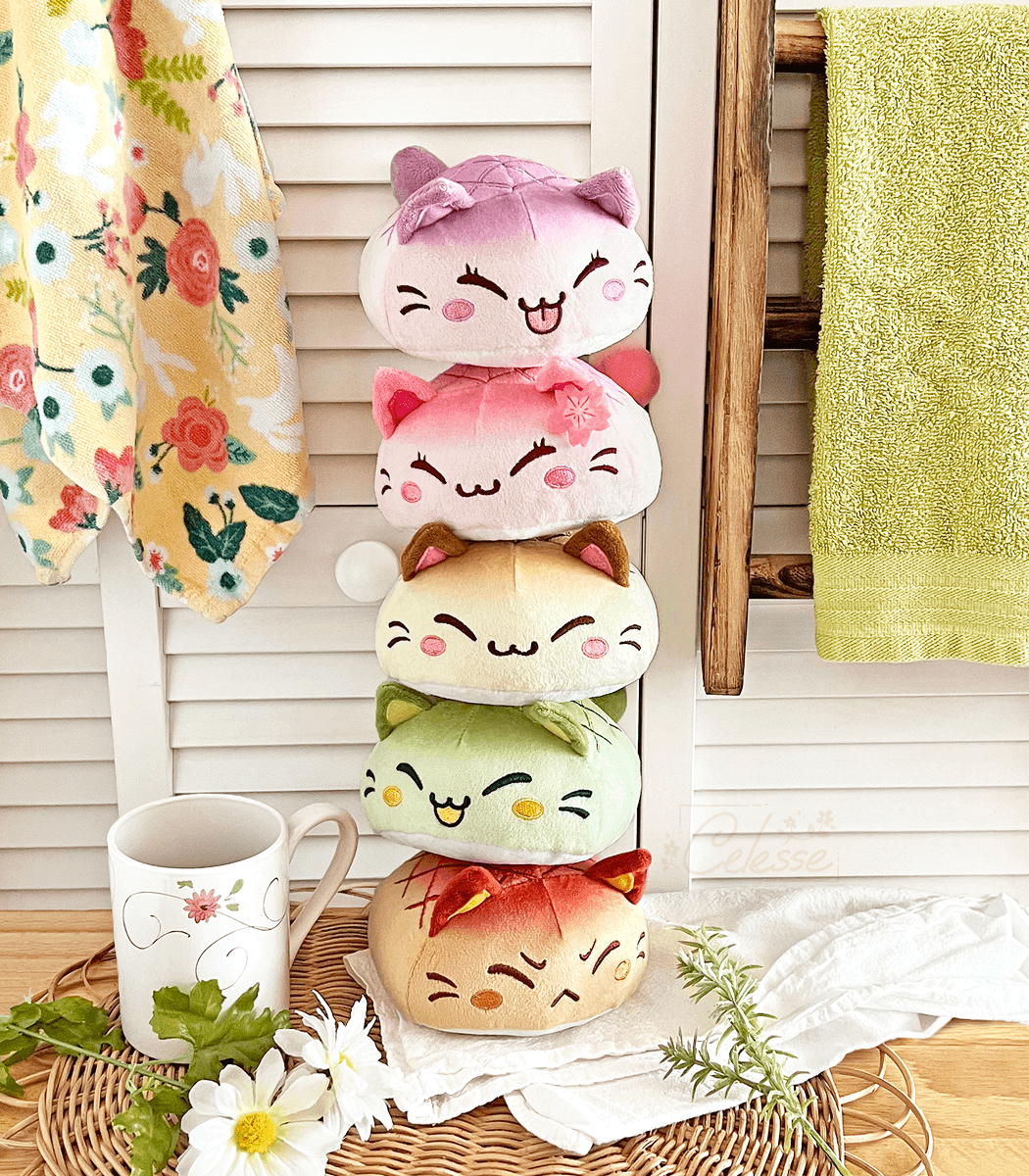 A photo of a vertical stack of plush of bread cats in various "flavors" on a kitchen counter. From the top down they are Taro, Sakura, Original, Matcha and Red Bean. They each have a squinty cat face and are colored to match their flavors.