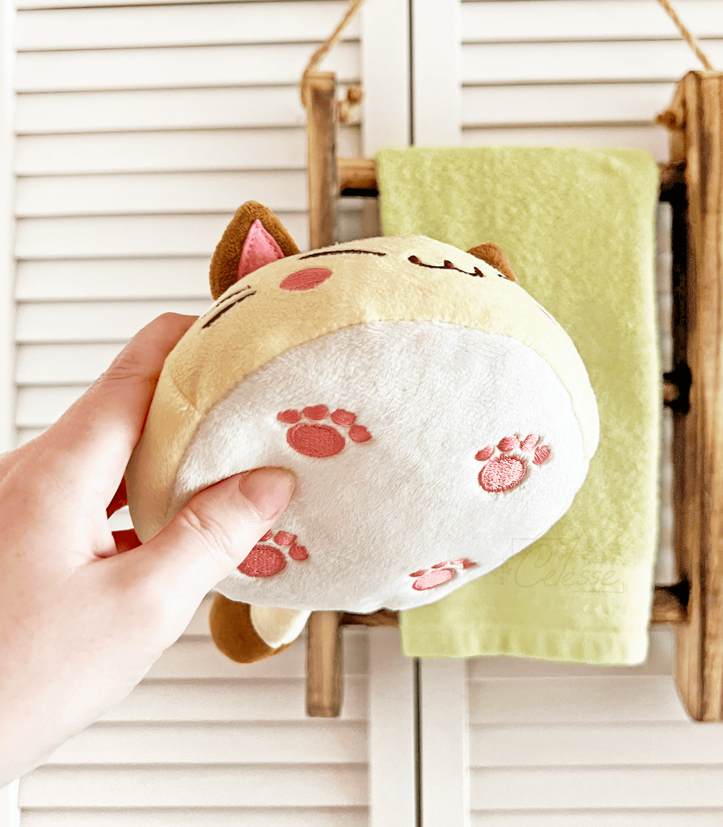 A photo of a hand holding a round bread cat plush, showing off the underside where little paws are embroidered on the bottom.