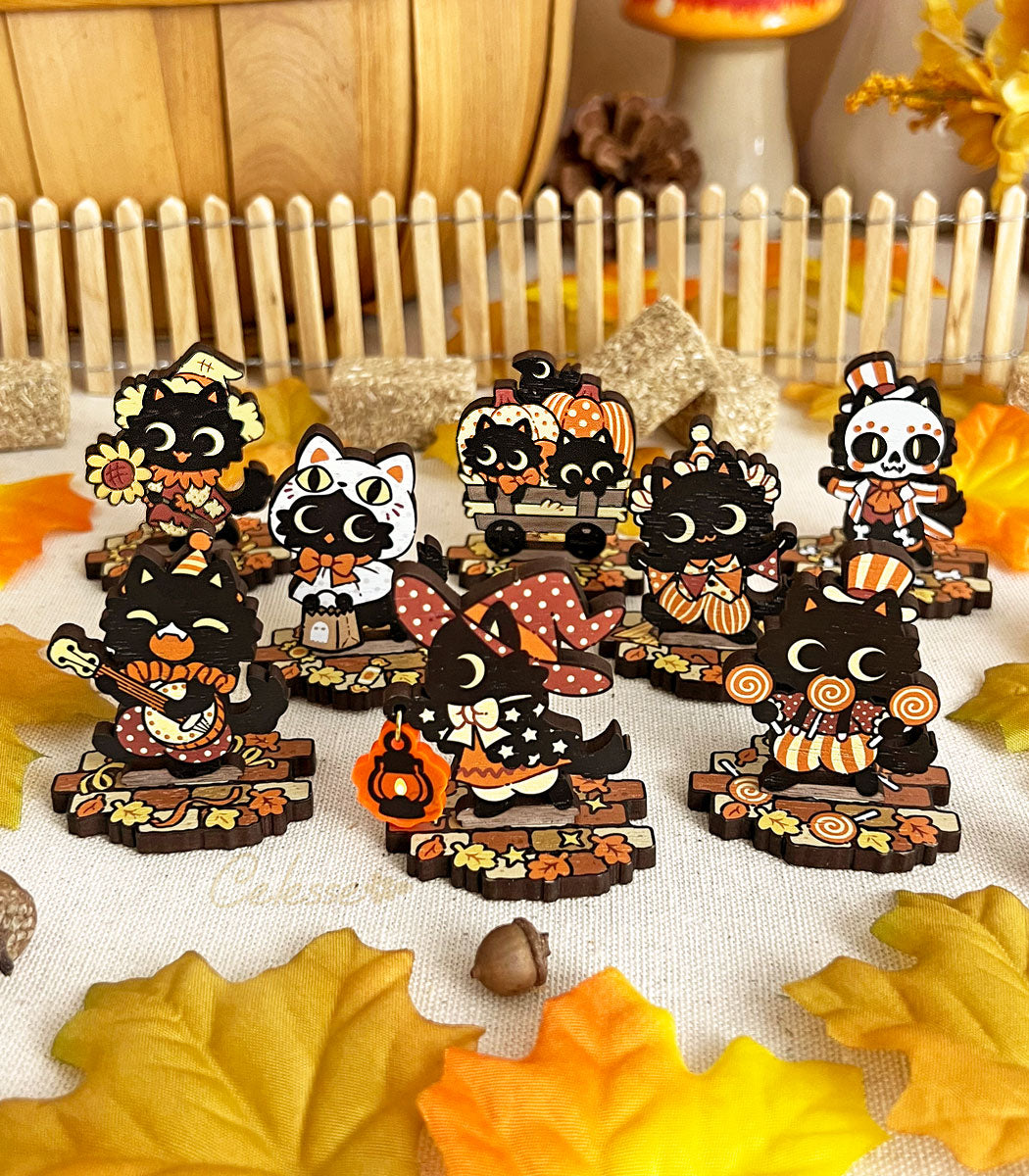 A collection of wooden standees depicting whimsical black cat characters in Halloween attire, arranged on a surface with autumn leaves and a picket fence.