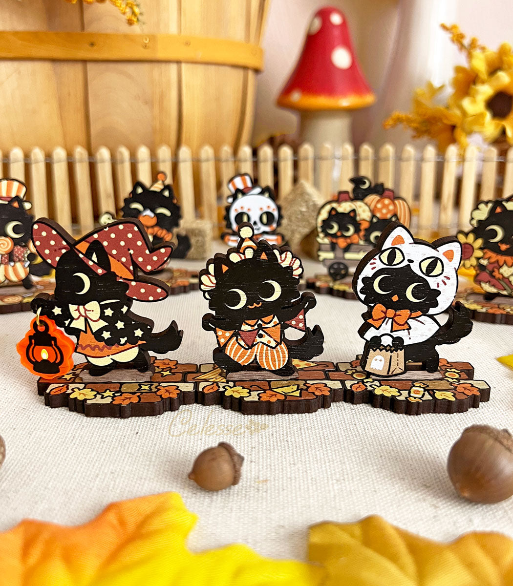 A collection of wooden standees depicting whimsical black cat characters in Halloween attire, arranged on a surface with autumn leaves and a picket fence.