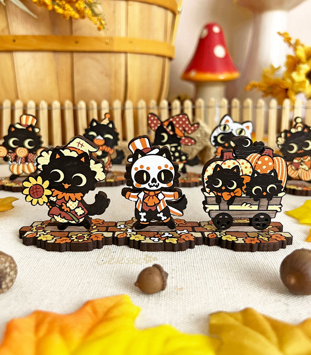 A collection of wooden standees depicting whimsical black cat characters in Halloween attire, arranged on a surface with autumn leaves and a picket fence.
