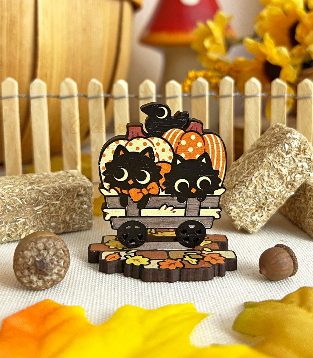 A wooden standee depicting whimsical black cat characters in Halloween attire, arranged on a surface with autumn leaves and a picket fence.
