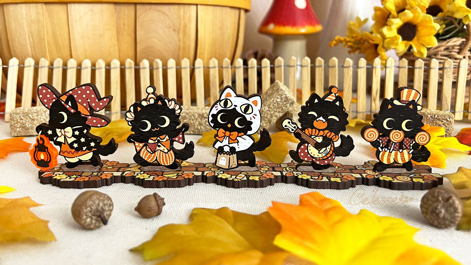 A collection of wooden standees depicting whimsical black cat characters in Halloween attire, arranged on a surface with autumn leaves and a picket fence.