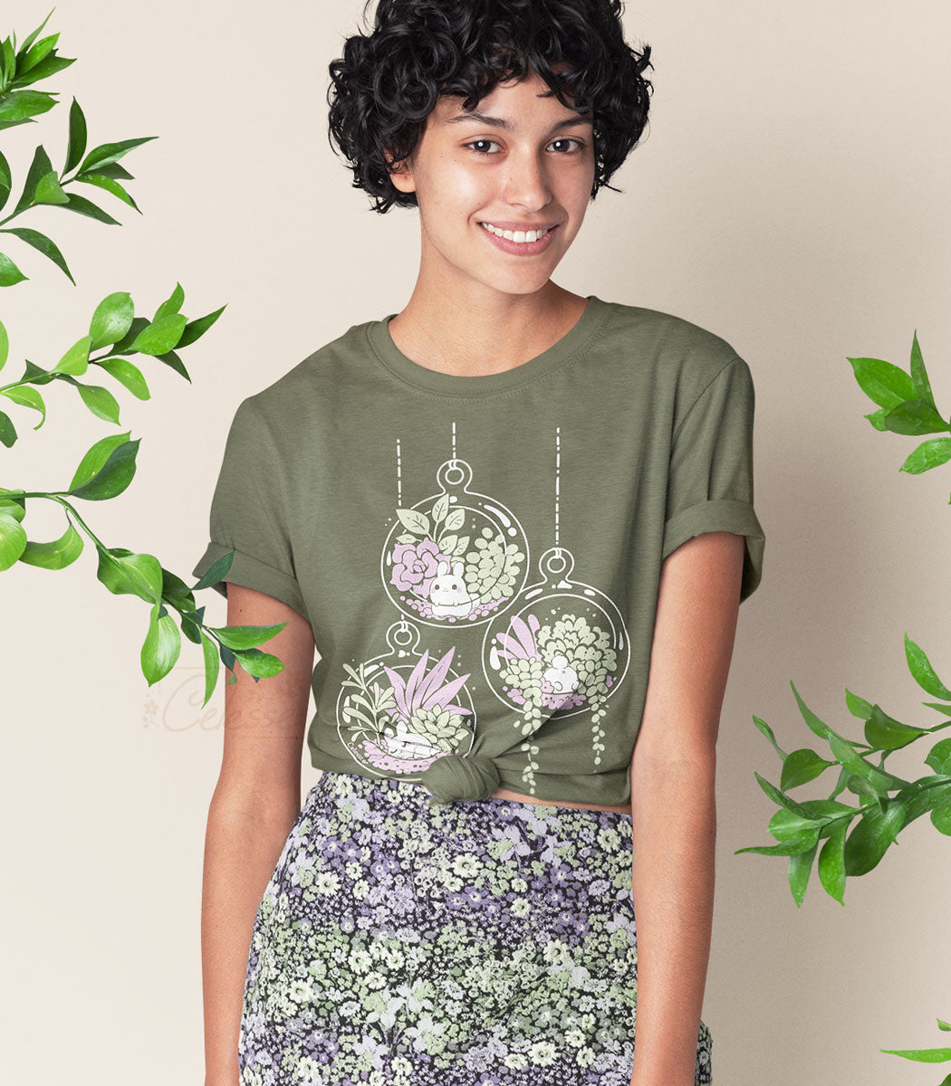 A photo of a dark-haired woman wearing a light olive shirt printed with terrarium globes filled with plants and each having a tiny bunny inside them. Leaves surround her.