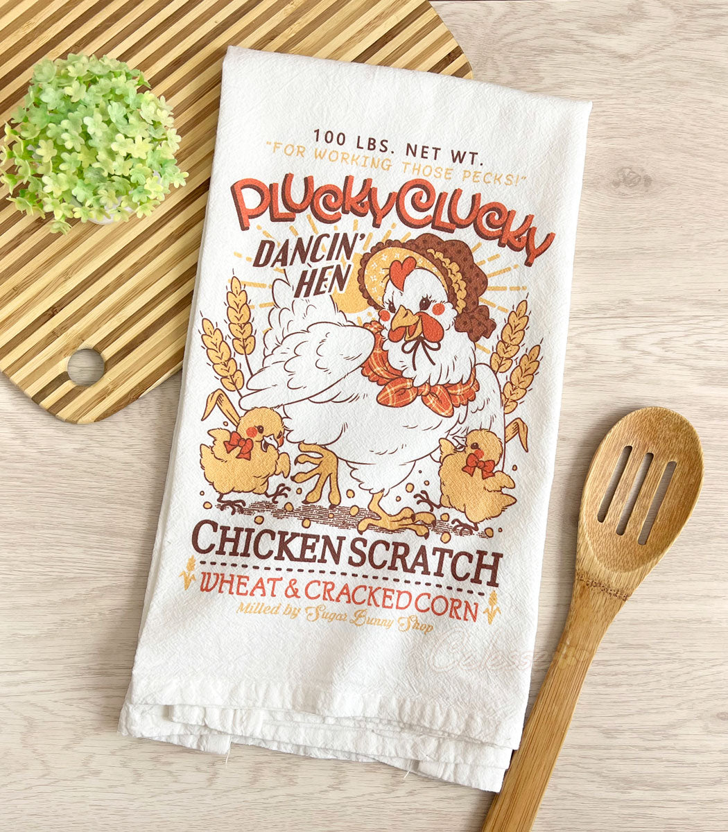 A photo of a white flour sack tea towel laid on a table, a cutting board and plant in the top left corner, a slotted wooden spoon in the bottom right. The towel print features a faux vintage-style logo for Plucky Clucky Chicken Scratch with a dancing hen in a bonnet and two chicks