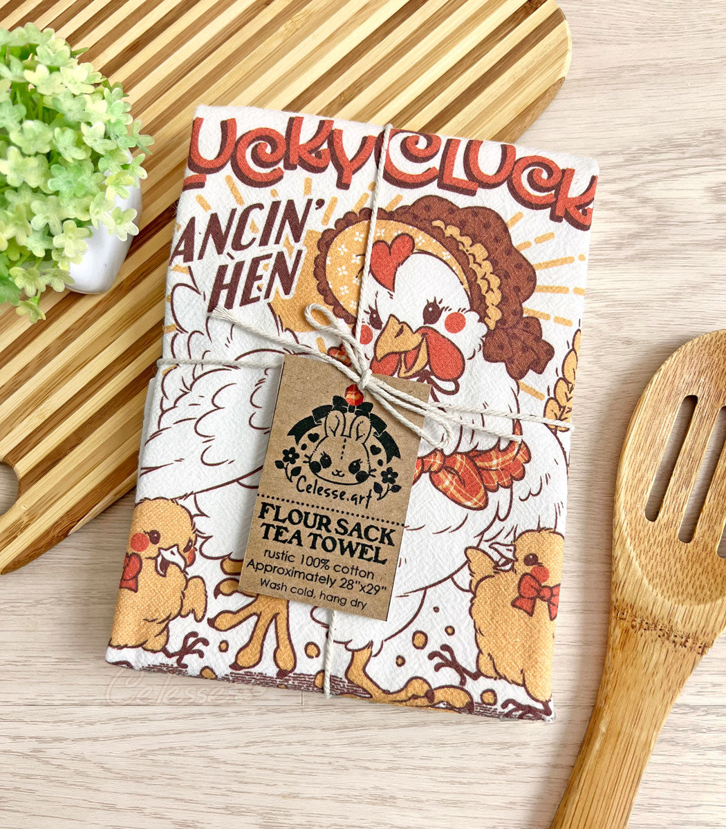 A photo of a white flour sack tea towel laid on a table, a cutting board and plant in the top left corner, a slotted wooden spoon in the bottom right. The towel print features a faux vintage-style logo for Plucky Clucky Chicken Scratch with a dancing hen in a bonnet and two chicks