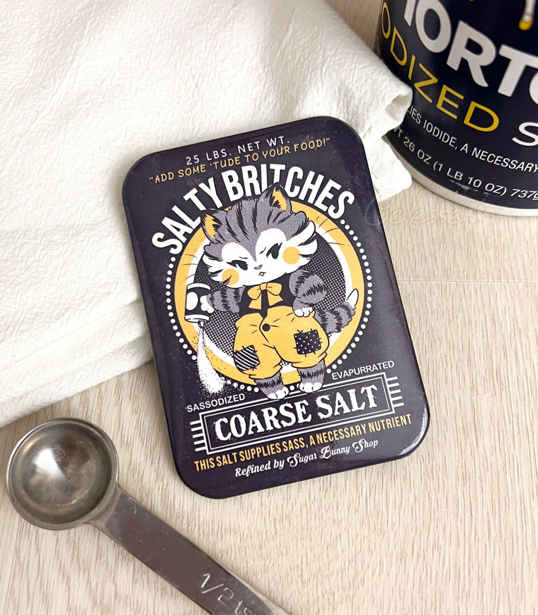 A photo of a fridge magnet with artwork featuring a vintage-style logo for Salty Britches Coarse Salt with a kitten wearing patchwork britches and petulantly pouring out a container of salt.