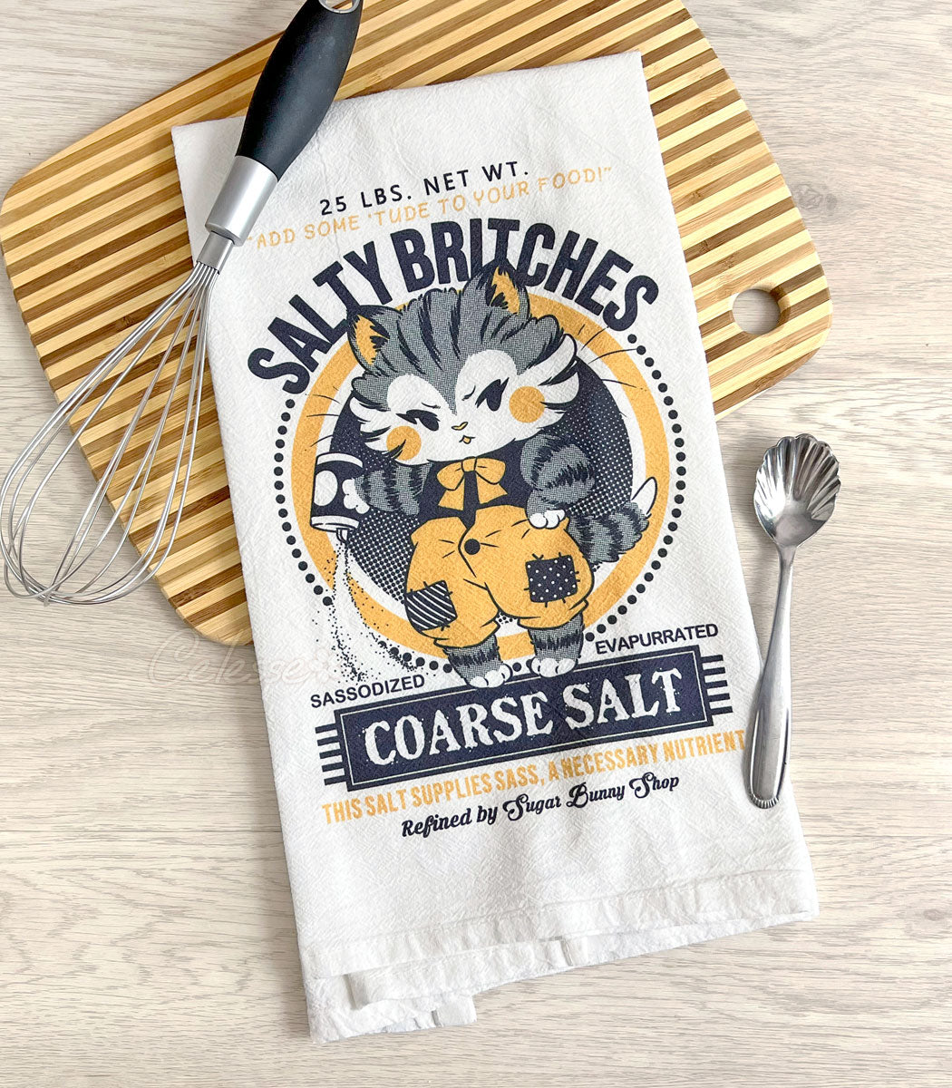 A white tea towel on a kitchen counter with a striped cutting board and some utensils, the printed design featuring a vintage-style logo for Salty Britches Coarse Salt with a kitten wearing patchwork britches and petulantly pouring out a container of salt.
