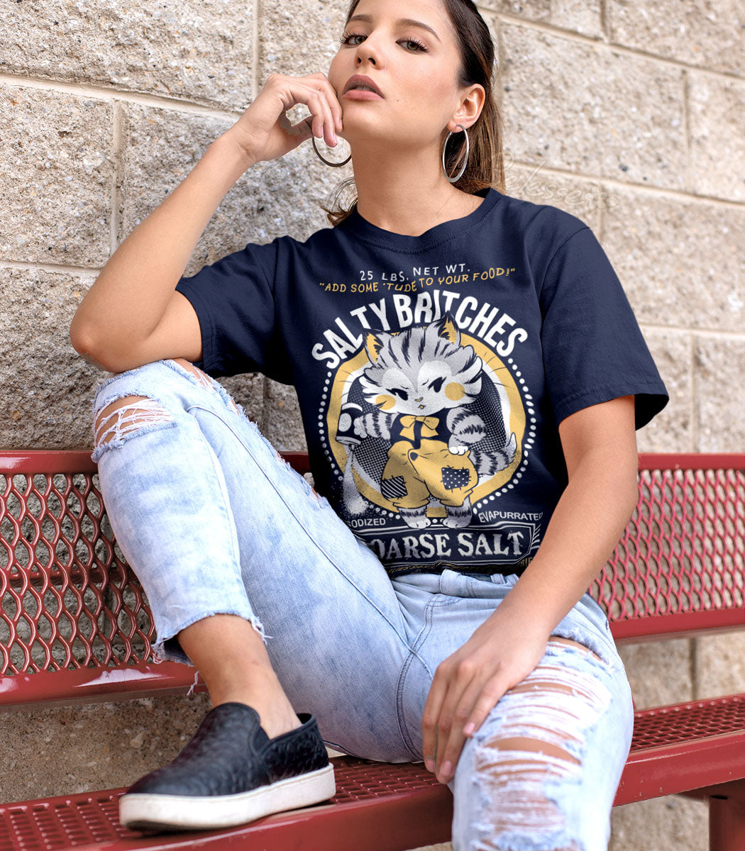A young woman sitting on a bench with her foot up on the seat and elbow rested on it, wearing a navy blue shirt with a faux logo for Salty Britches Coarse Salt. The logo features a grumpy kitten dumping a container of salt in a petulant manner