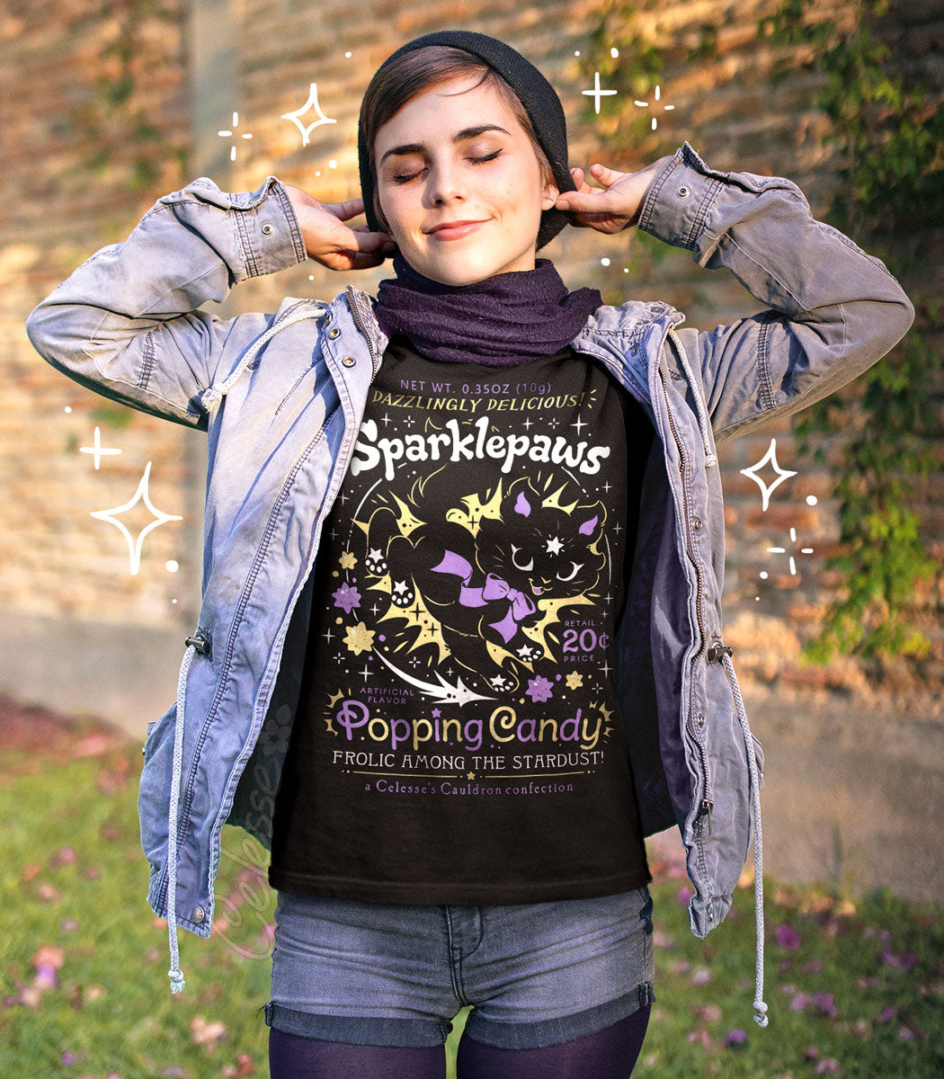A woman wearing a black shirt with a print of a faux logo for Sparklepaws Popping Candy with a magical cat, styled with an open light-wash denim jacket and a dark scarf, posing outdoors against a blurred brick wall background with digital sparkle overlays.