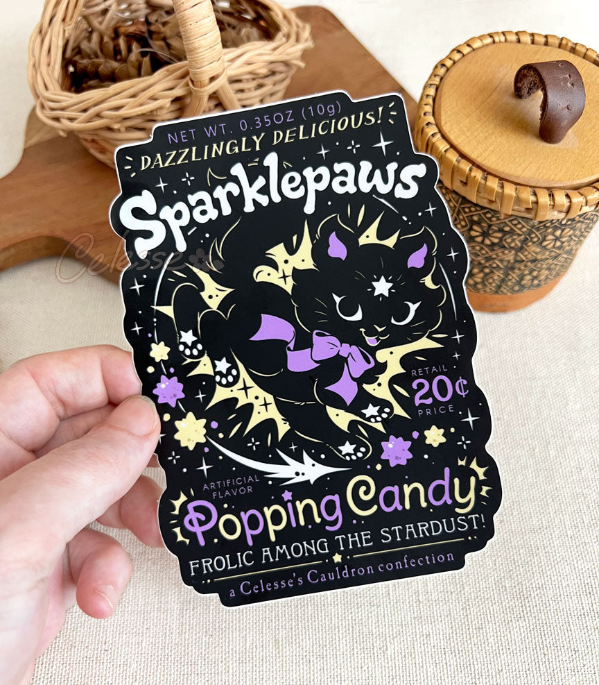 A hand holding a vinyl sticker with a print of a retro faux candy logo for Sparklepaws Popping Candy, the art featuring a black cat with star paw pads leaping among bursting sparkles. The flavor text reads, "Dazzlingly delicious!" and "Frolic among the stardust!"