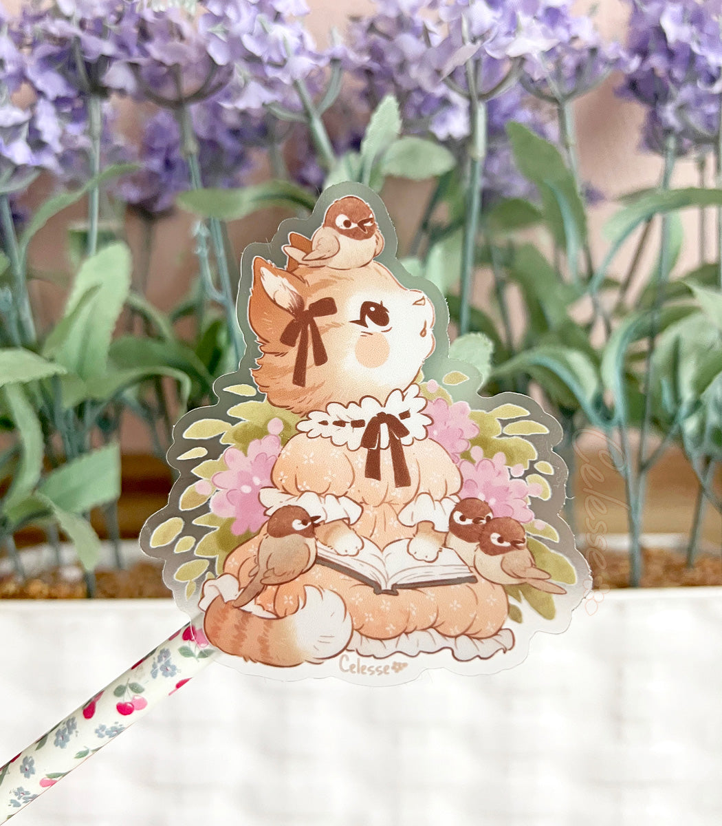 A photo of a clear vinyl sticker featuring artwork of a kitten reading a book in a vintage dress with birds, a basket of lavender flowers in the background.