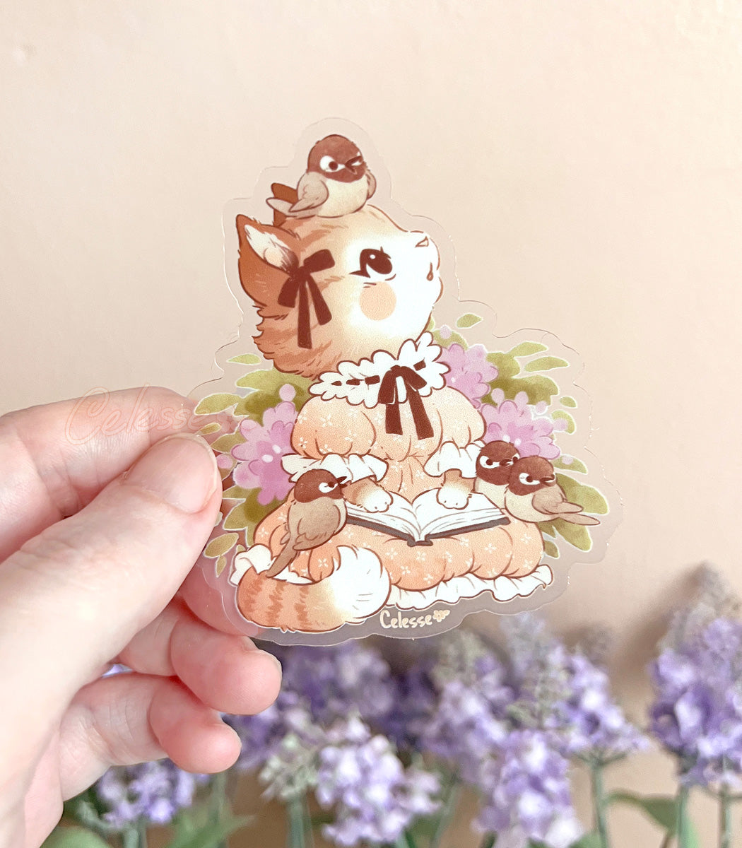 A photo of a hand holding a clear vinyl sticker of a cat reading a book with birds on a beige background