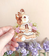 A photo of a hand holding a clear vinyl sticker of a cat reading a book with birds on a beige background
