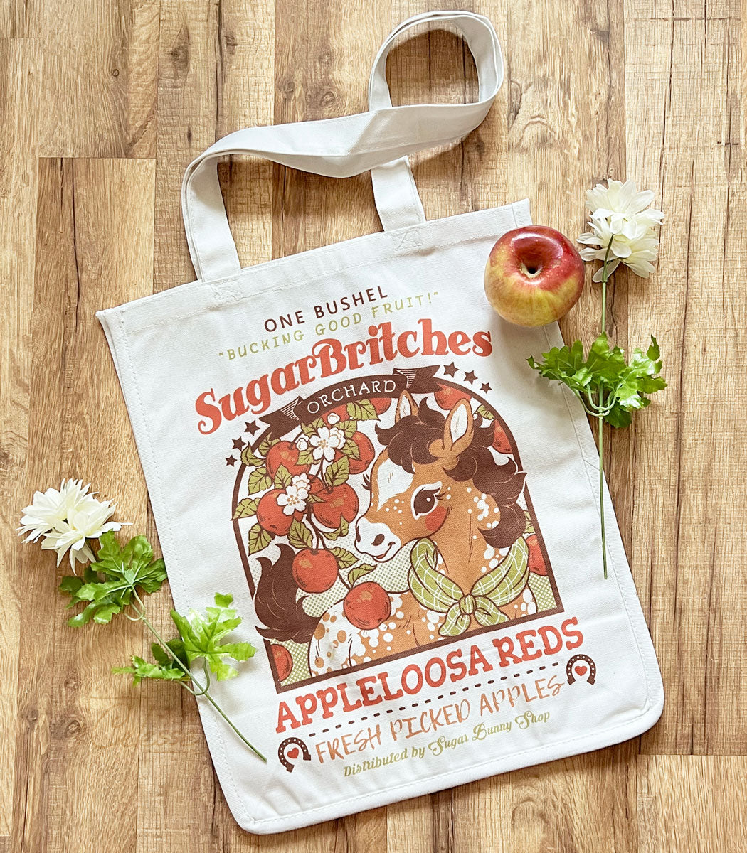 A photo of a white canvas tote bag laid on a wood table, white flowers and an apple surrounding it. The print is a faux vintage-style logo for SugarBritches Apple Orchard and features a cute baby horse with a branch of an apple tree.